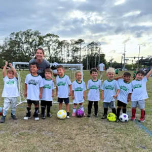 A group of children in matching shirts poses with a coach in front of a soccer goal, holding various soccer balls.