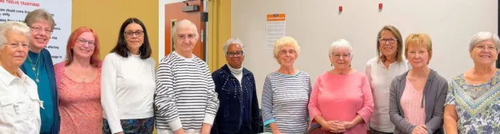 A group of women stands together, smiling, in a community space, with a poster visible in the background.