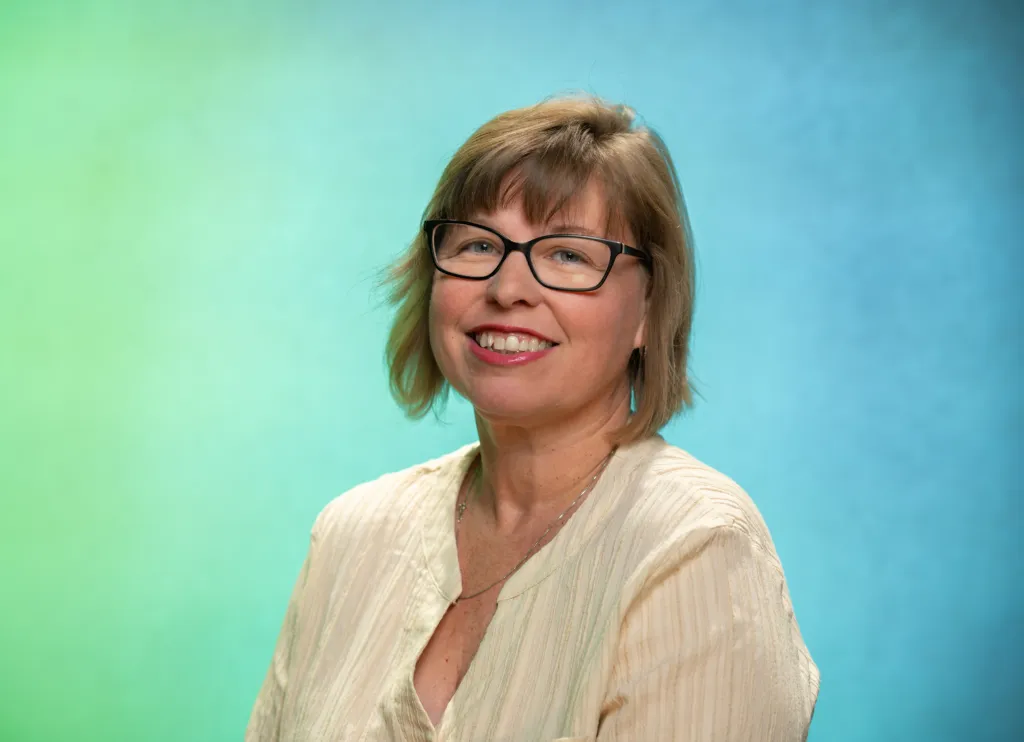 A woman with glasses smiles while posing against a gradient blue and green background.