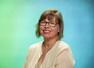 A woman with glasses smiles while posing against a gradient blue and green background.