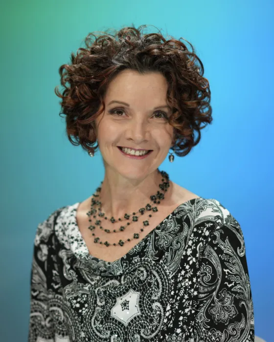 A woman with curly hair smiles while wearing a patterned top and necklace against a gradient background.