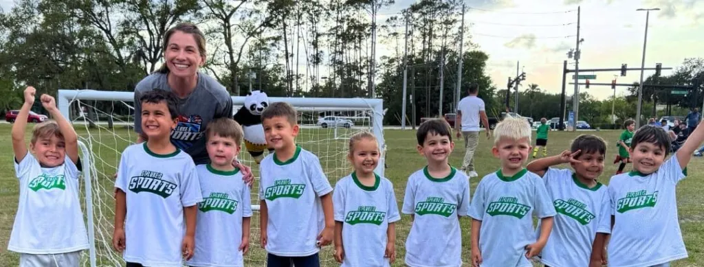 Children wearing matching shirts pose with a coach in front of a soccer goal, celebrating after a game.