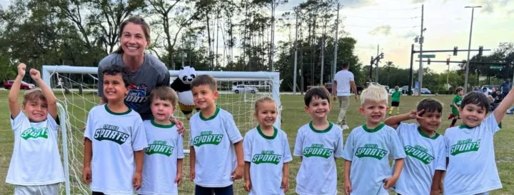 Children wearing matching shirts pose with a coach in front of a soccer goal, celebrating after a game.
