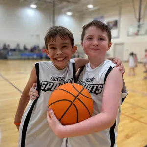 Two boys in sports jerseys smile while holding a basketball on a gym court.