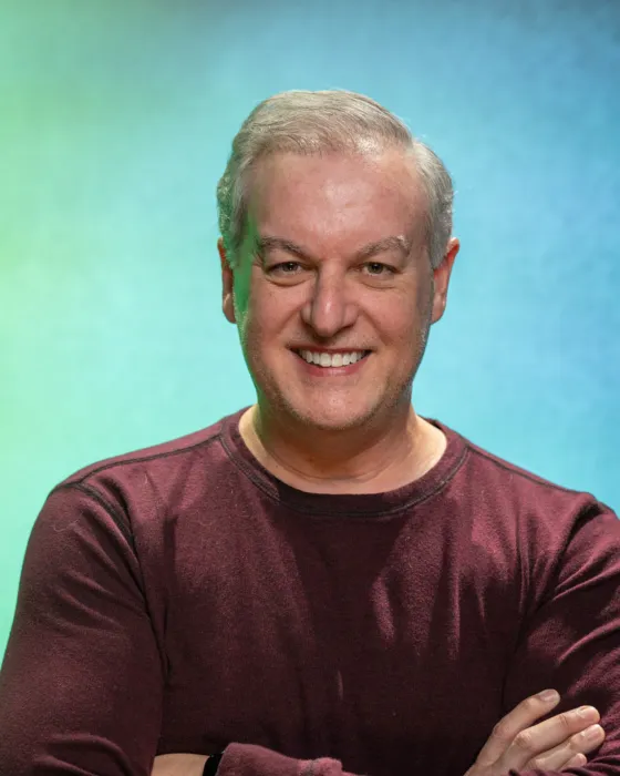 A man with short gray hair smiles while standing with arms crossed against a gradient blue and green background.