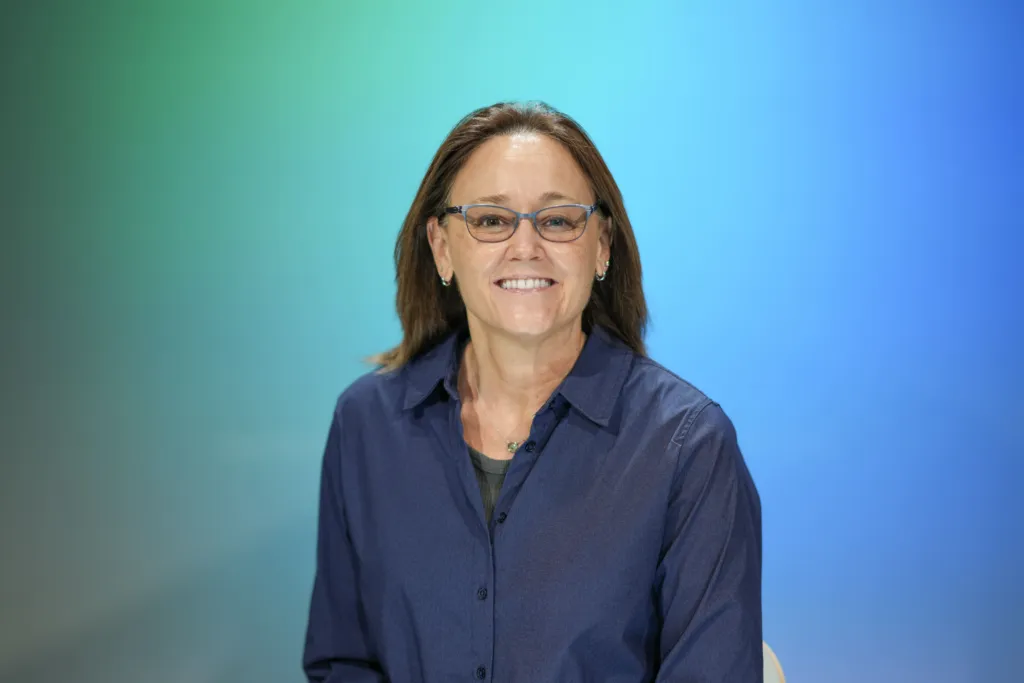 A woman with glasses smiles while seated against a gradient blue-green background.