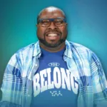 A man wearing glasses and a blue shirt that says "YOU BELONG" smiles while sitting against a gradient background.
