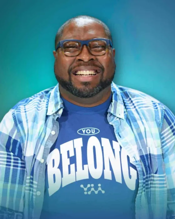 A man wearing glasses and a blue shirt that says "YOU BELONG" smiles while sitting against a gradient background.