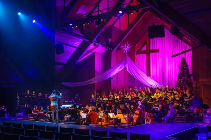 A choir performs on stage while musicians play instruments in a decorated church setting with colorful lighting.