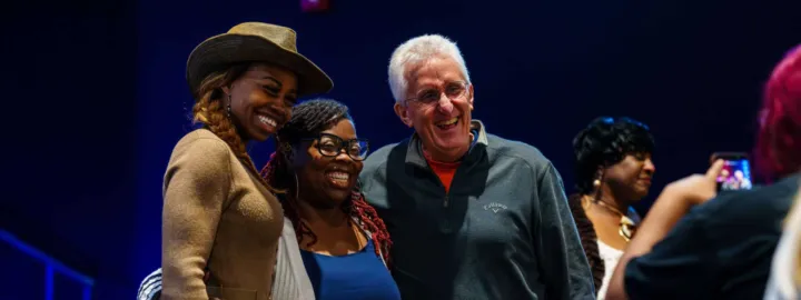 Three people pose for a photo together, smiling, while another person takes a picture in the background.
