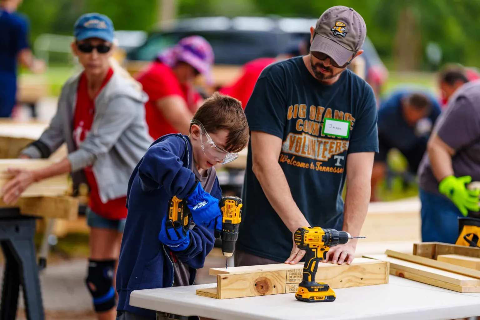 Children and adults are working together on a woodworking project, using drills and tools at a community event.