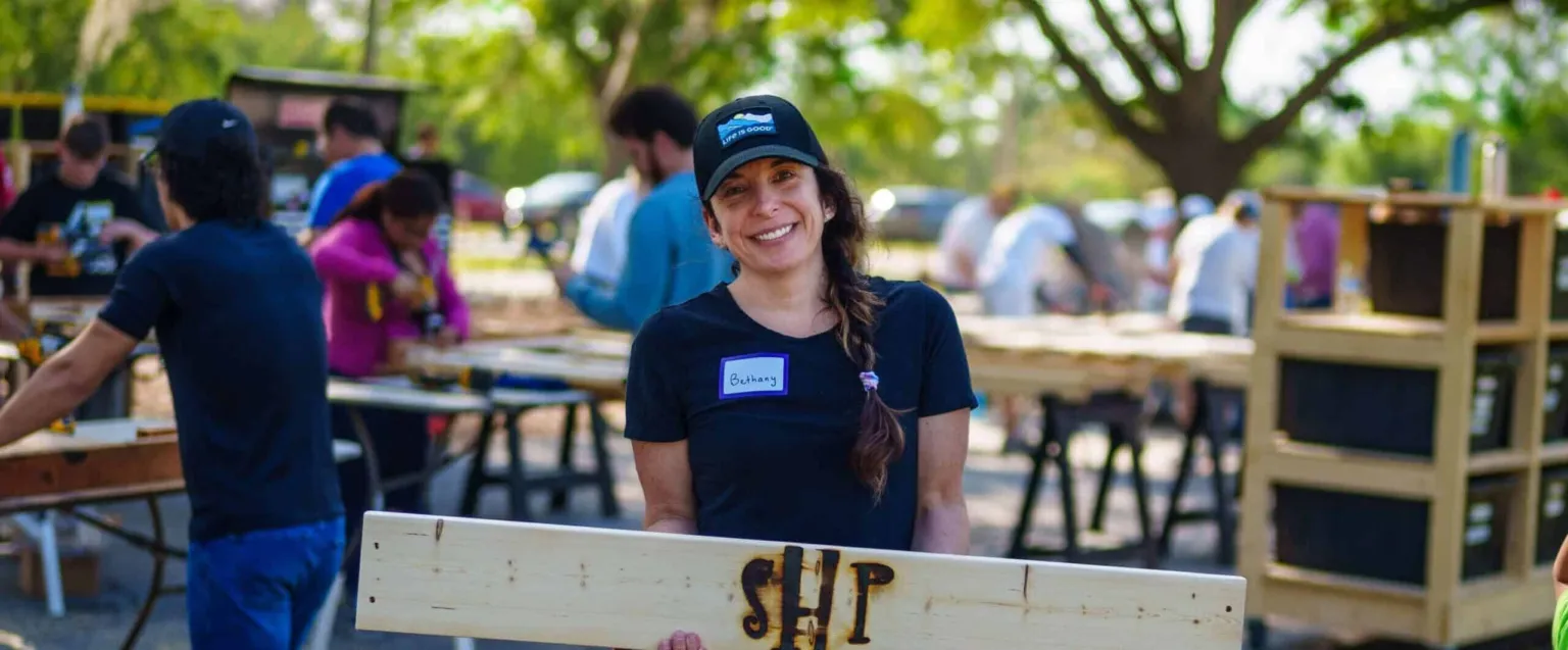 A volunteer holds a wooden sign while working on a community project with others in the background.