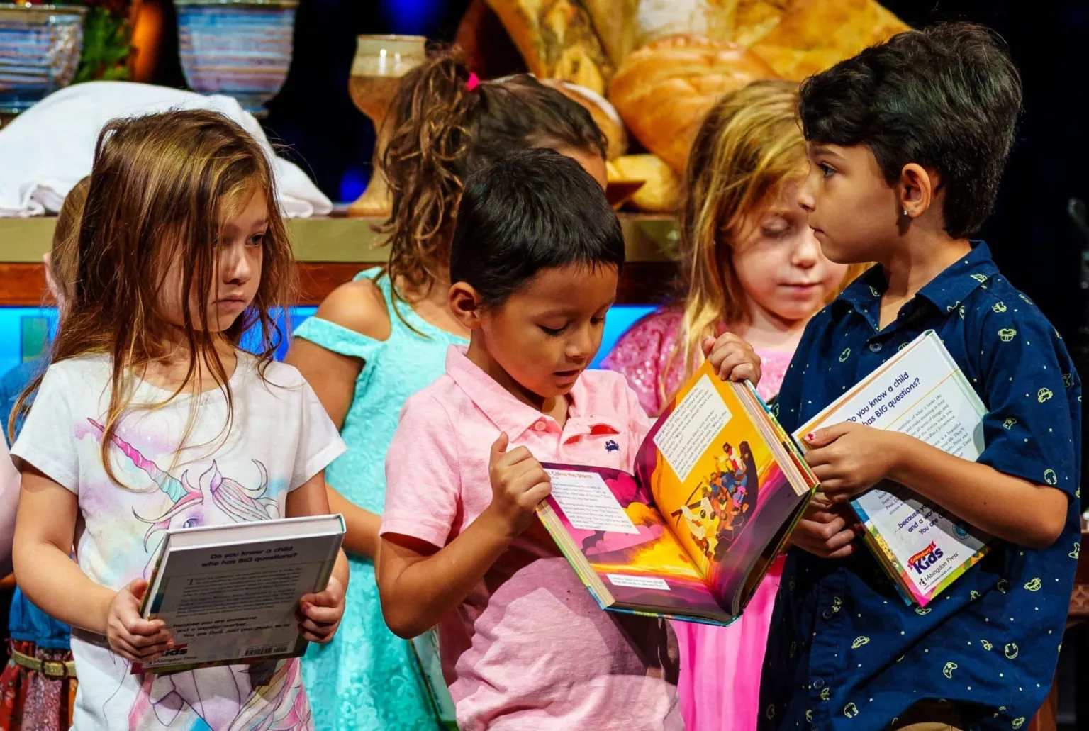 Children stand together, holding books and looking at each other while engaging in a group activity.