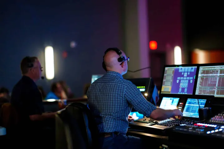 Technicians operate sound and lighting equipment at a church event, focusing on multiple computer screens.