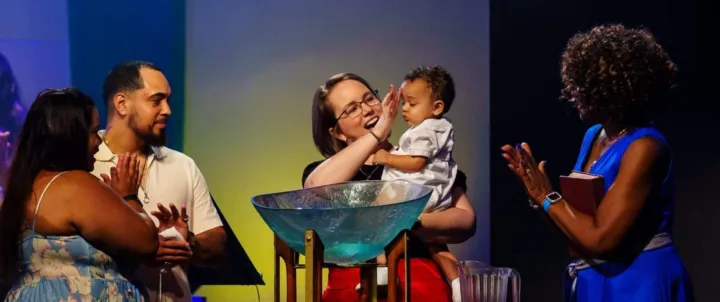 A woman holds a baby above a baptismal font while others applaud during a church ceremony.