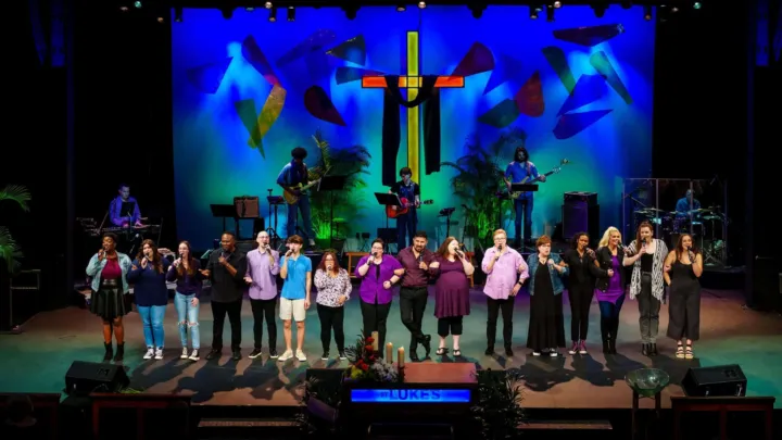 A diverse group of singers performs on stage in front of a colorful backdrop with a cross.