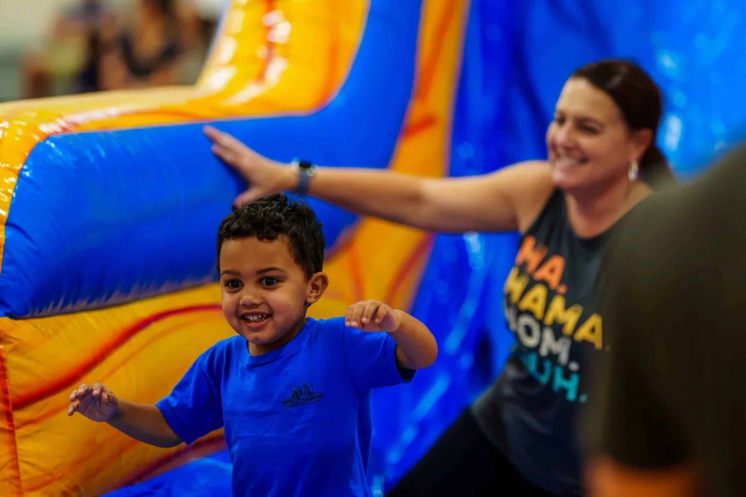 A child in a blue shirt smiles while playing near a colorful inflatable structure, with an adult nearby.