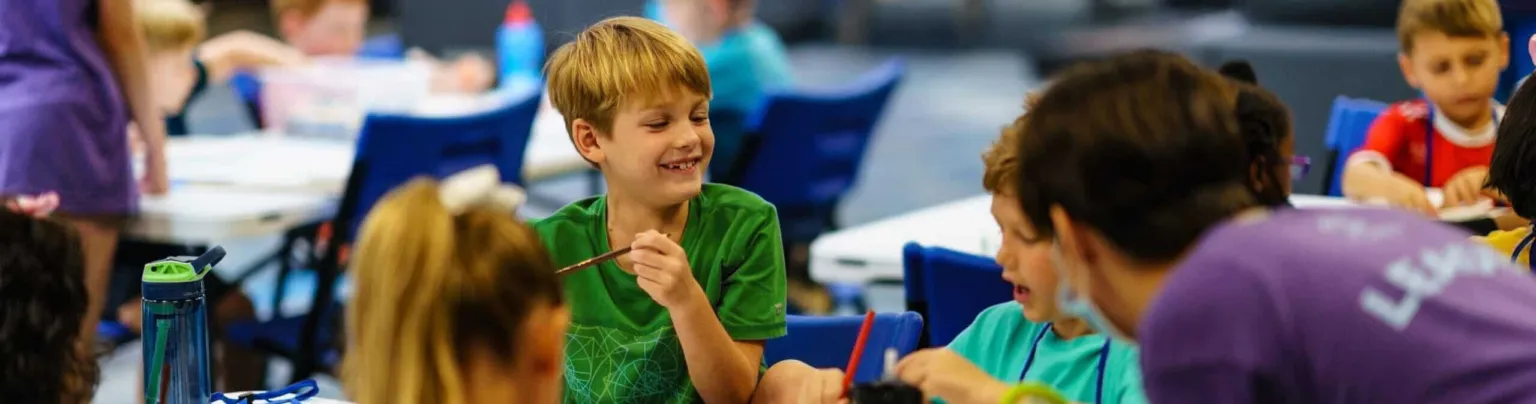 Children are engaged in art activities at tables, smiling and interacting with each other and an adult leader.