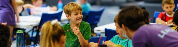 Children are engaged in art activities at tables, smiling and interacting with each other and an adult leader.
