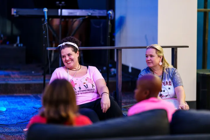 Two women sit on a couch, engaging in conversation, while a child in a pink shirt is partially visible in the foreground.