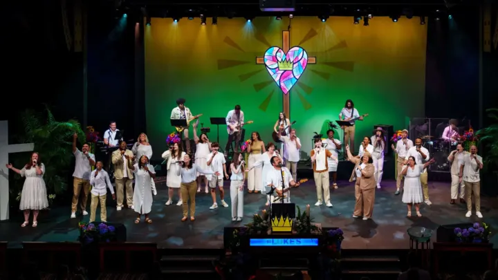 A large group of singers and musicians perform on stage, dressed in white, with a colorful stained glass heart backdrop.
