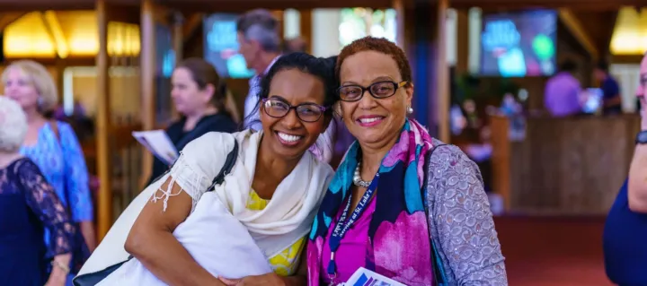 Two women smile and pose for a photo while holding pamphlets in a busy church gathering space.