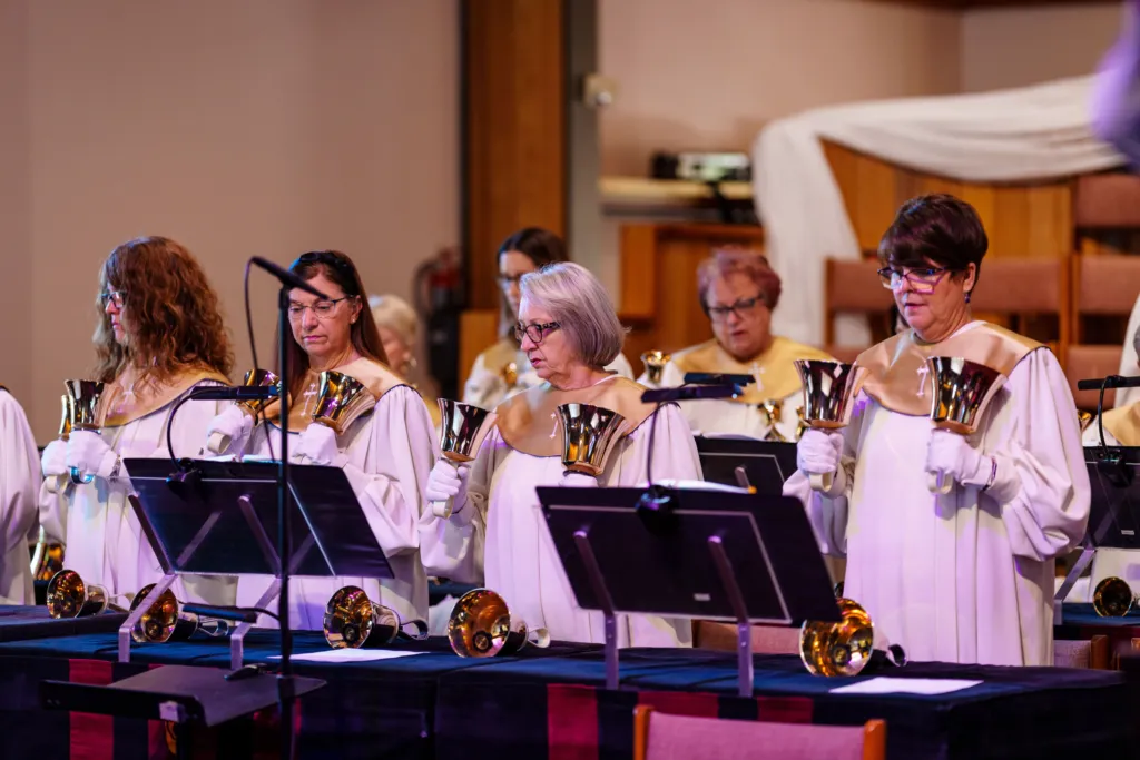 A handbell choir performs, holding bells and focusing on their music stands during a church service.