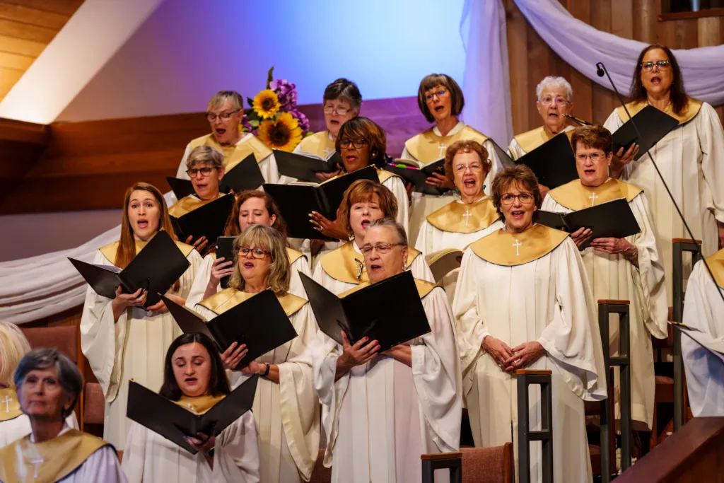 A choir of women in white robes with gold accents sings together on a stage, holding music sheets.