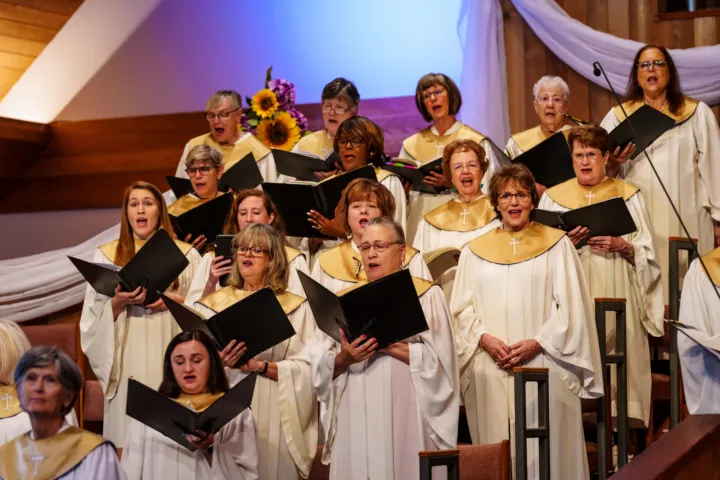 A choir of women in white robes with gold accents sings together on a stage, holding music sheets.