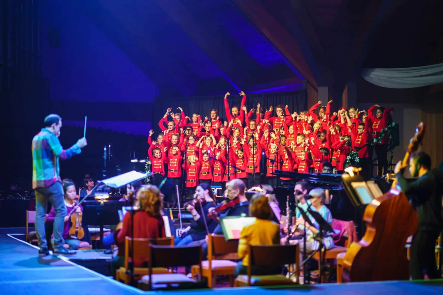 A conductor leads a children's choir in red outfits while musicians play instruments on stage.