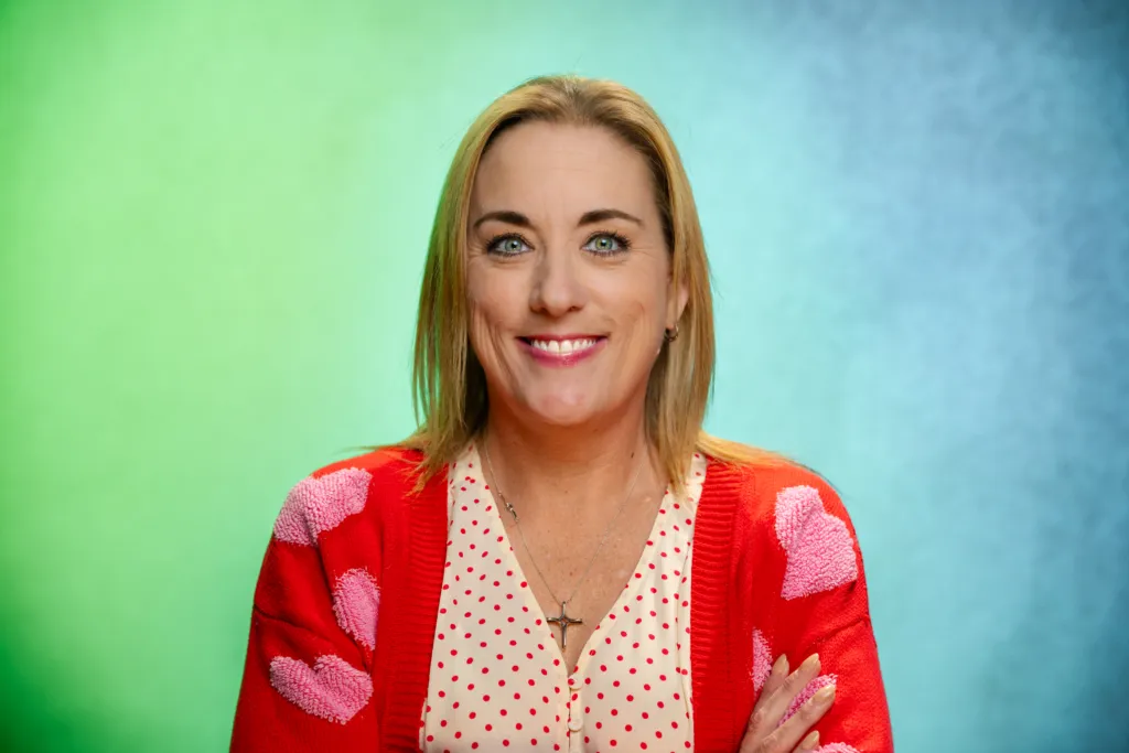 A woman with blonde hair smiles while wearing a red cardigan with heart patterns over a polka dot top.
