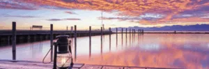 A lantern sits on a dock, reflecting on calm water under a colorful sunset sky with clouds.