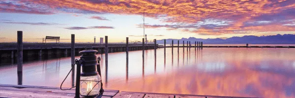 A lantern sits on a dock, reflecting on calm water under a colorful sunset sky with clouds.