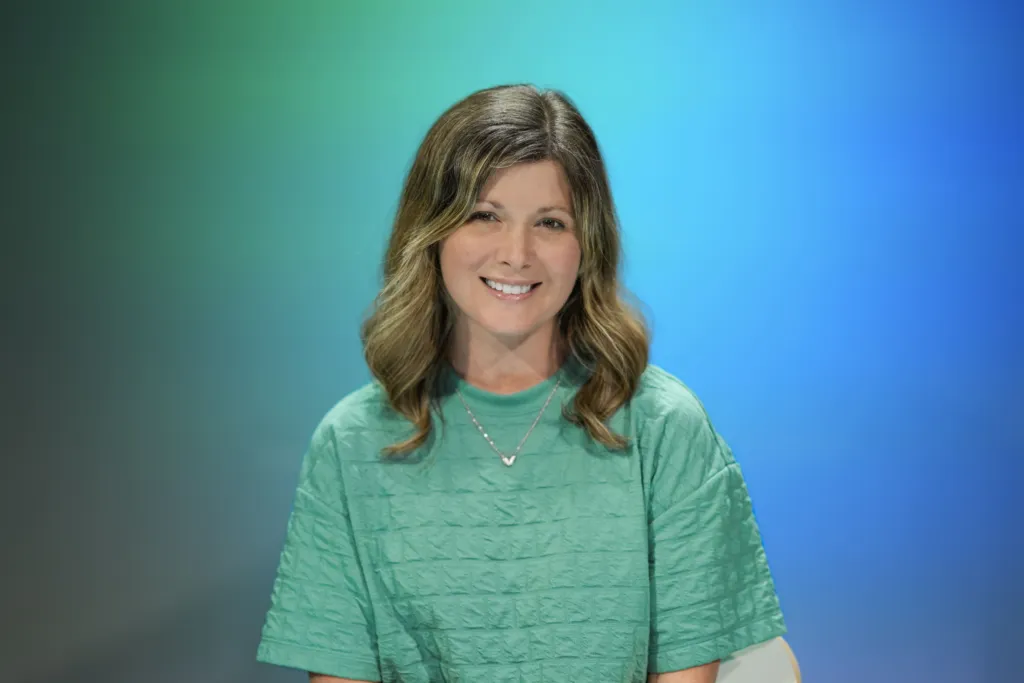 A woman with wavy hair smiles while wearing a green top against a gradient blue background.
