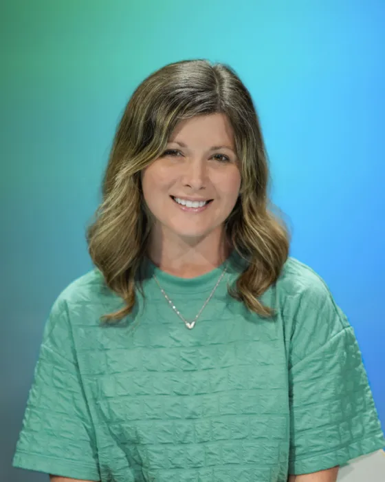 A woman with wavy hair smiles while wearing a green top against a gradient blue background.