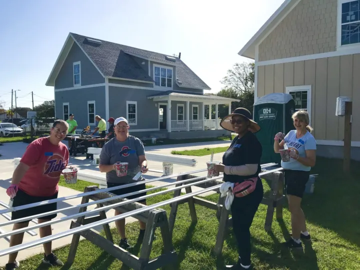Volunteers paint wooden beams outdoors, with a house and portable restroom in the background.
