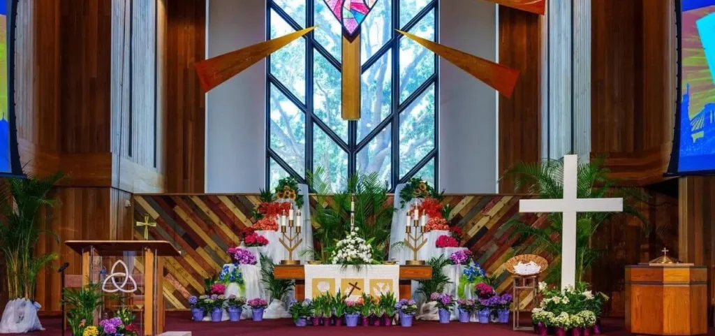 The altar is decorated with flowers and candles, featuring a large white cross and a colorful stained glass window behind it.