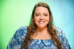 A woman with long, wavy hair smiles while wearing a blue floral dress against a gradient green and blue background.