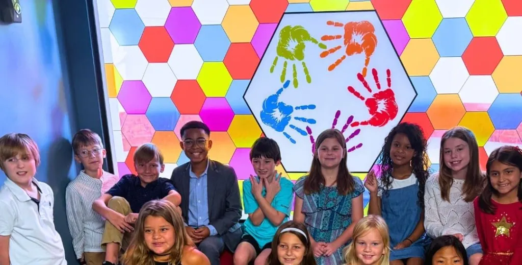 A group of children poses together in front of a colorful hexagonal backdrop featuring handprints.