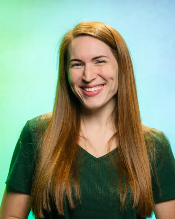 A woman with long hair smiles while standing against a gradient green and blue background.