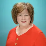 A woman with short, wavy hair smiles while wearing a bright orange top against a teal background.
