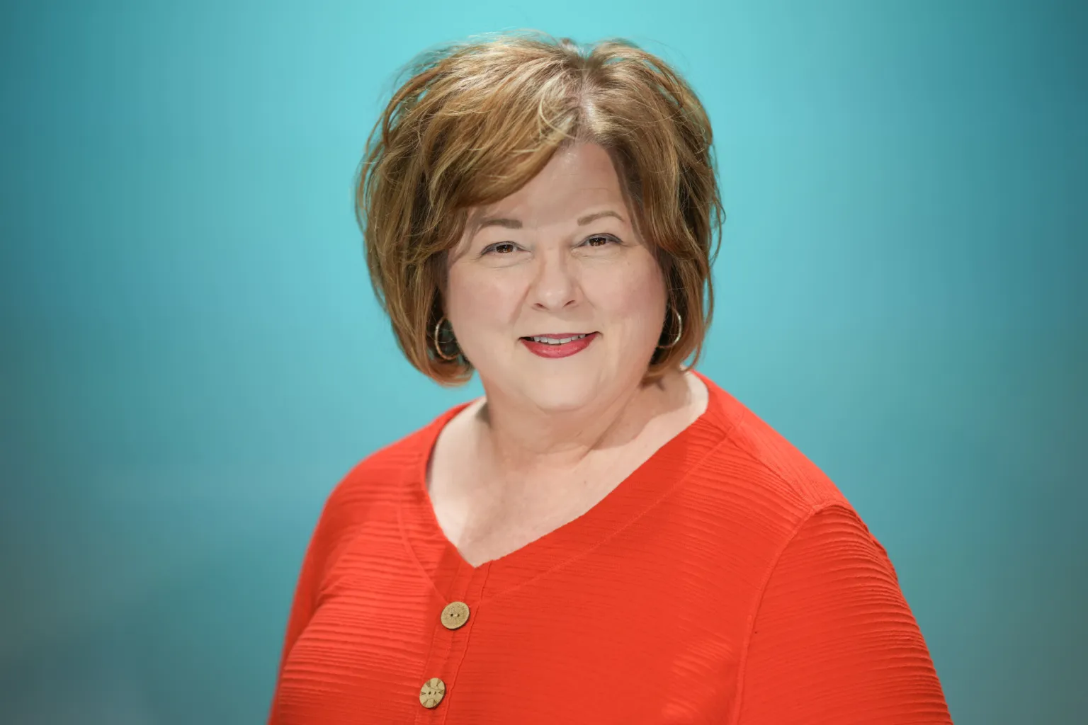 A woman with short, wavy hair smiles while wearing a bright orange top against a teal background.