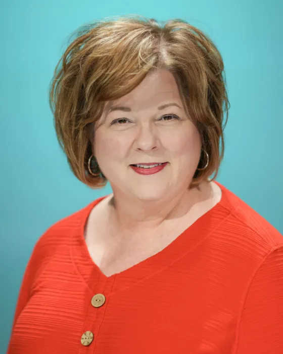 A woman with short, wavy hair smiles while wearing a bright orange top against a teal background.