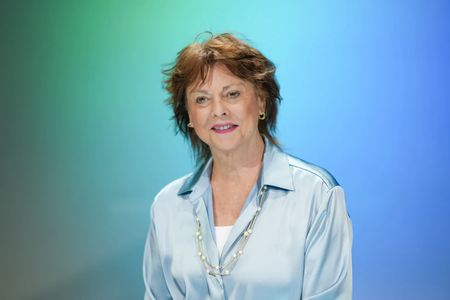 A woman with short, tousled hair smiles while wearing a light blue blouse against a gradient background.