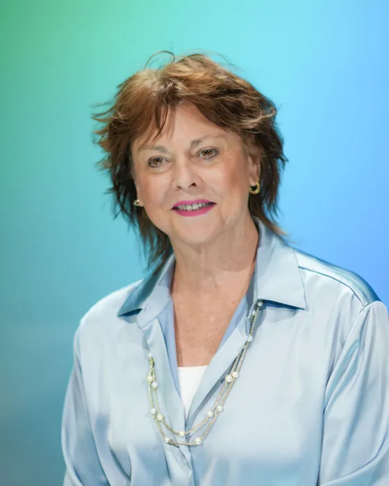 A woman with short, tousled hair smiles while wearing a light blue blouse against a gradient background.