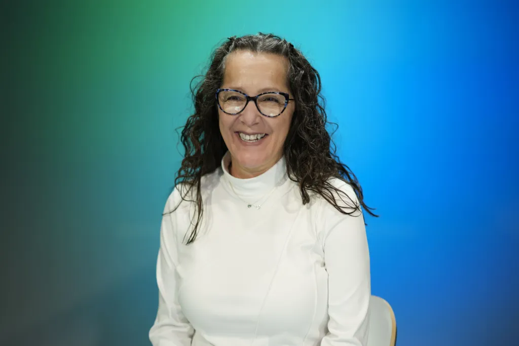 A woman with curly hair and glasses smiles while seated against a blue and green gradient background.