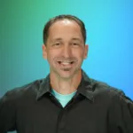 A man smiles while standing against a gradient blue background, wearing a black shirt and light blue undershirt.
