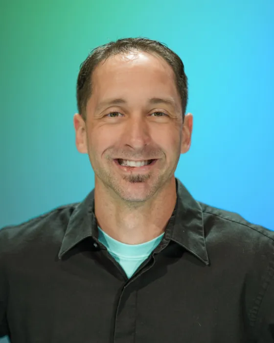 A man smiles while standing against a gradient blue background, wearing a black shirt and light blue undershirt.