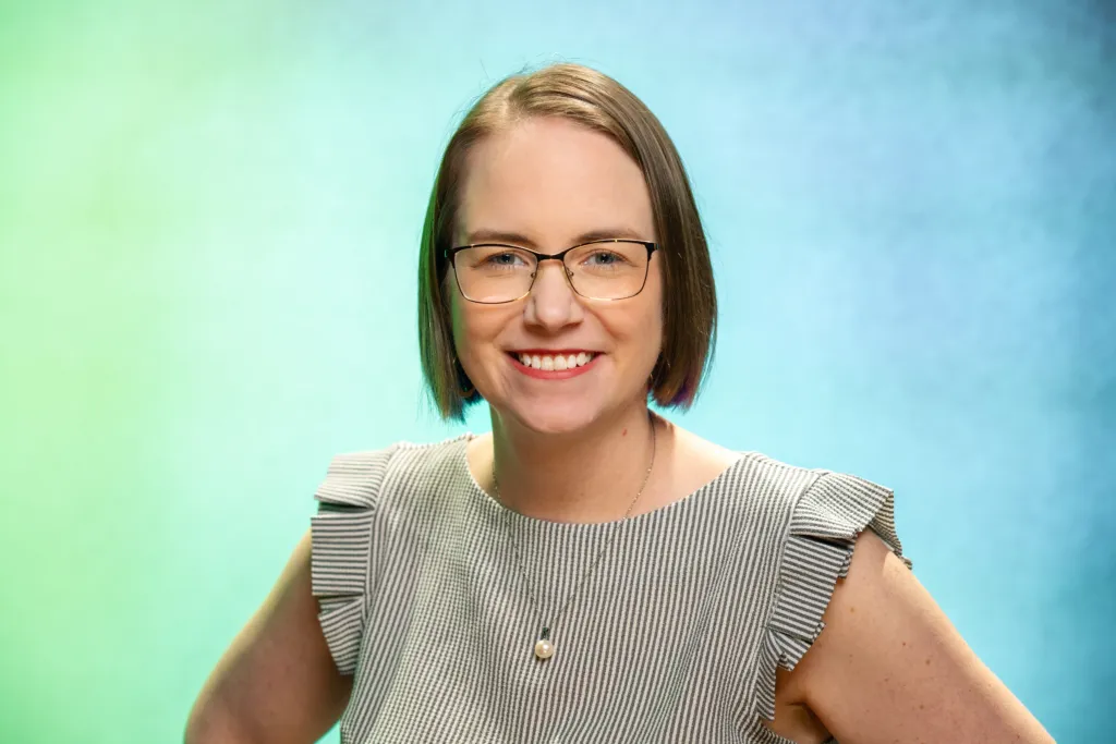 A woman with short hair and glasses smiles while wearing a striped top against a blue and green background.