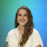 A woman with curly hair smiles while wearing a white shirt against a blue and green background.